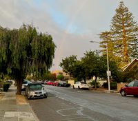 A residential San Mateo street lined with parked cars on both curbs, with a faint rainbow in the sky above the rooftops